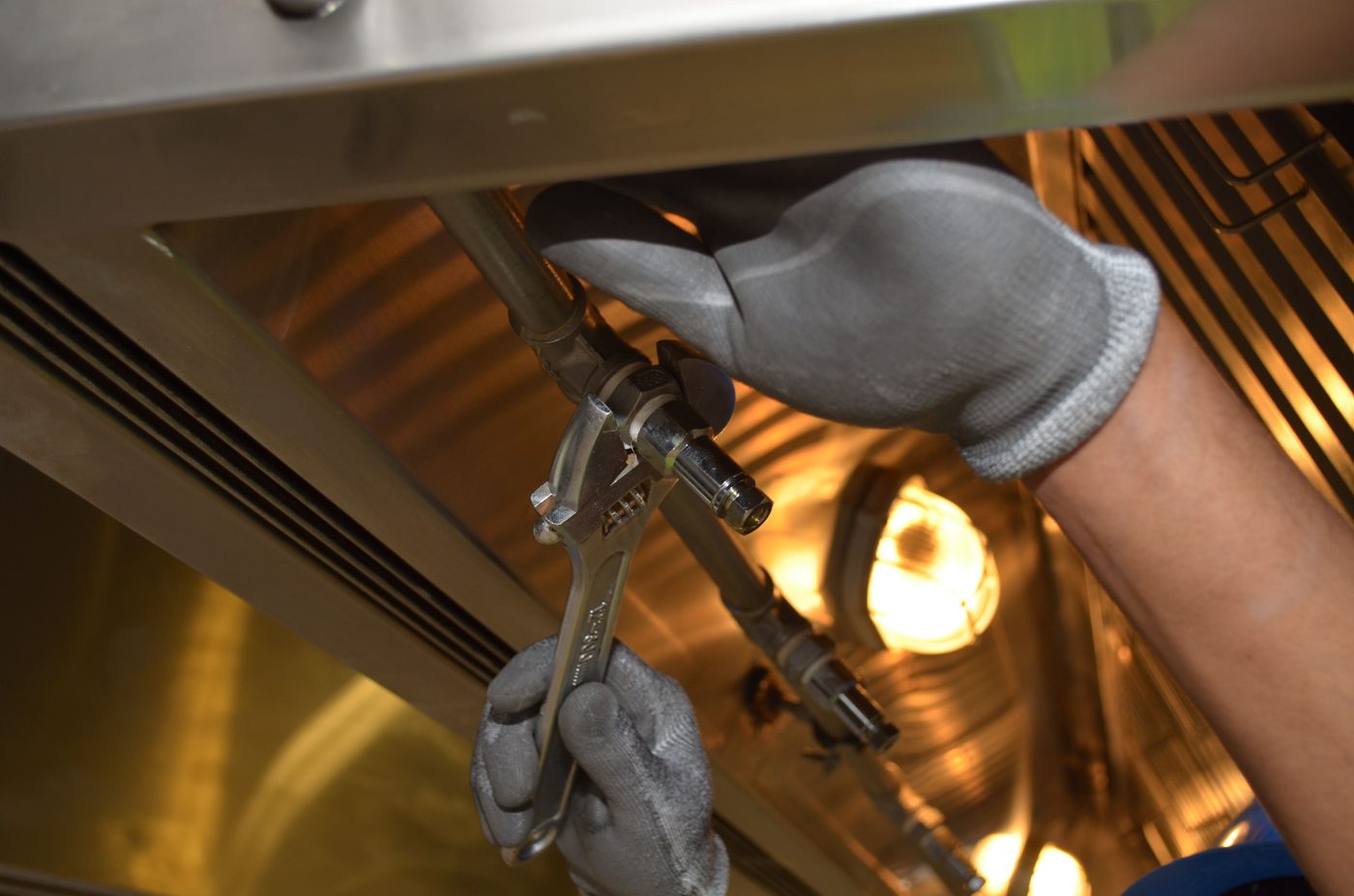 Gloved hands working a wrench on pipework under a warm work light